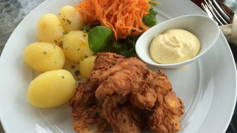 Stegt fisk med kartofler, revet gulerod, salat og mayonnaise på en hvid tallerken.