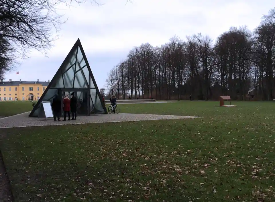 Triangulært glashus i parkområde, indgang til underjordisk museum. Udstillingsgenstande i mørkt rum med belysning, inklusiv skulptur og installationer.