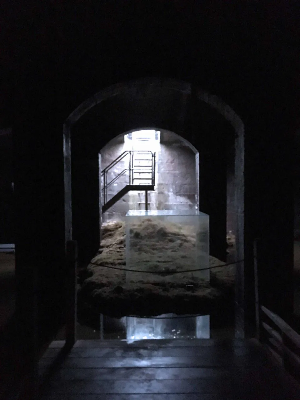 Triangulært glashus i parkområde, indgang til underjordisk museum. Udstillingsgenstande i mørkt rum med belysning, inklusiv skulptur og installationer.