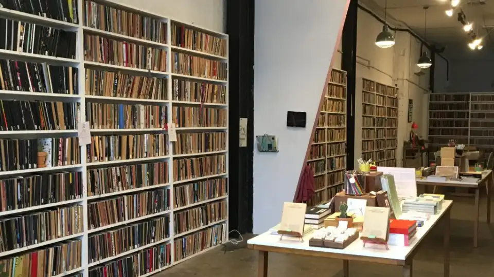 Bibliotek eller boghandel med hylder fyldt med bøger og borde med forskellige skriveartikler.