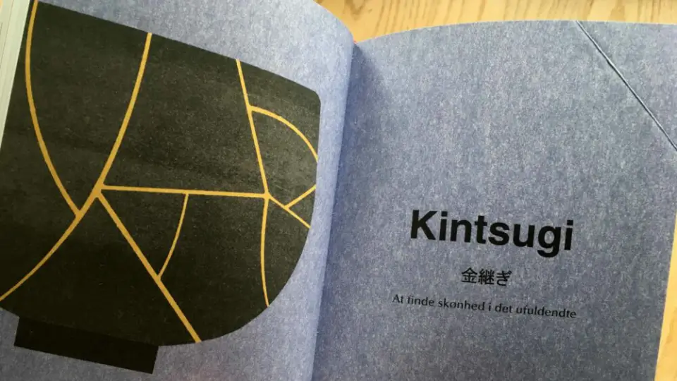 En åben bogside med ordet "Kintsugi" og en illustration af en sort skål repareret med guldlinjer. Teksten under forklarer konceptet "At finde skønhed i det ufuldendte