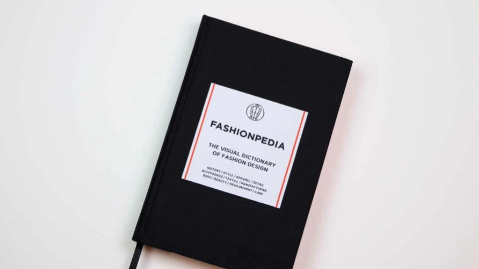 Bogen "Fashionpedia: The Visual Dictionary of Fashion Design" i sort hardcover på en hvid baggrund.