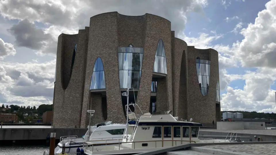 Moderne bygning med unikke buede former og glasvinduer, set fra havnen med både i forgrunden.