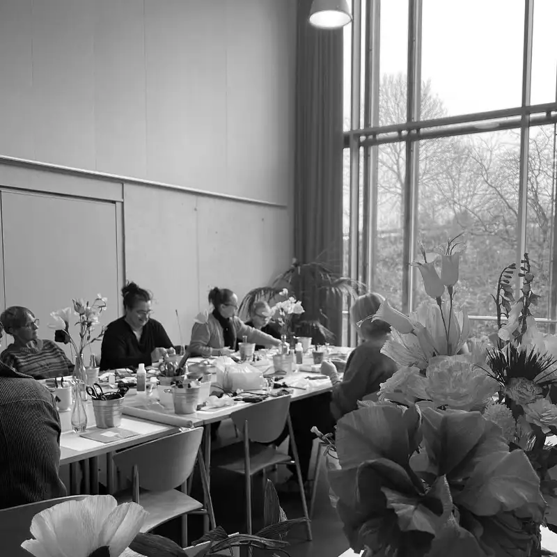 Personer sidder ved et bord og arbejder med papirblomster i et lokale med store vinduer.