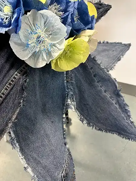 Billedet viser en kunstnerisk blomst sammensat af denim kronblade og farverige crepepapirblomster, arrangeret i en iøjnefaldende og kreativ komposition.